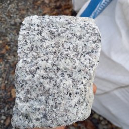 F.H.U. RAFBRUK RAFAŁ BŁAZY - Szary blok kostki granitowej o nieregularnych krawędziach, trzymany w dłoni, z widoczną strukturą kamienia i tłem z drobnym żwirem oraz workiem budowlanym.
