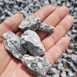 F.H.U. RAFBRUK RAFAŁ BŁAZY - Dłoń trzymająca kilka bryłek szarej kostki granitowej na tle wysypanego żwiru. Widoczne drobne użylenia i struktura kamienia.