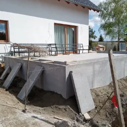 Betonowy taras w trakcie budowy przy białym domu z ciemnym dachem. Widoczne podpory i przygotowany teren. W tle drzewo i błękitne niebo.