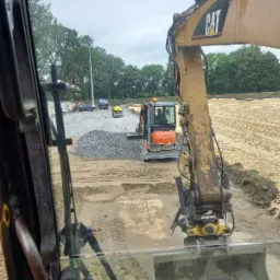 Widok z kabiny na prace ziemne: mini koparka Kubota wysypuje żwir, walec drogowy ubija teren. W tle zaparkowany samochód ciężarowy i przygotowany teren pod budowę.