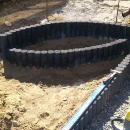 Formowanie rabaty z obrzeży betonowych imitujących drewno, poziomica na krawędzi, częściowo wypełniona ziemią, w tle kostka brukowa.