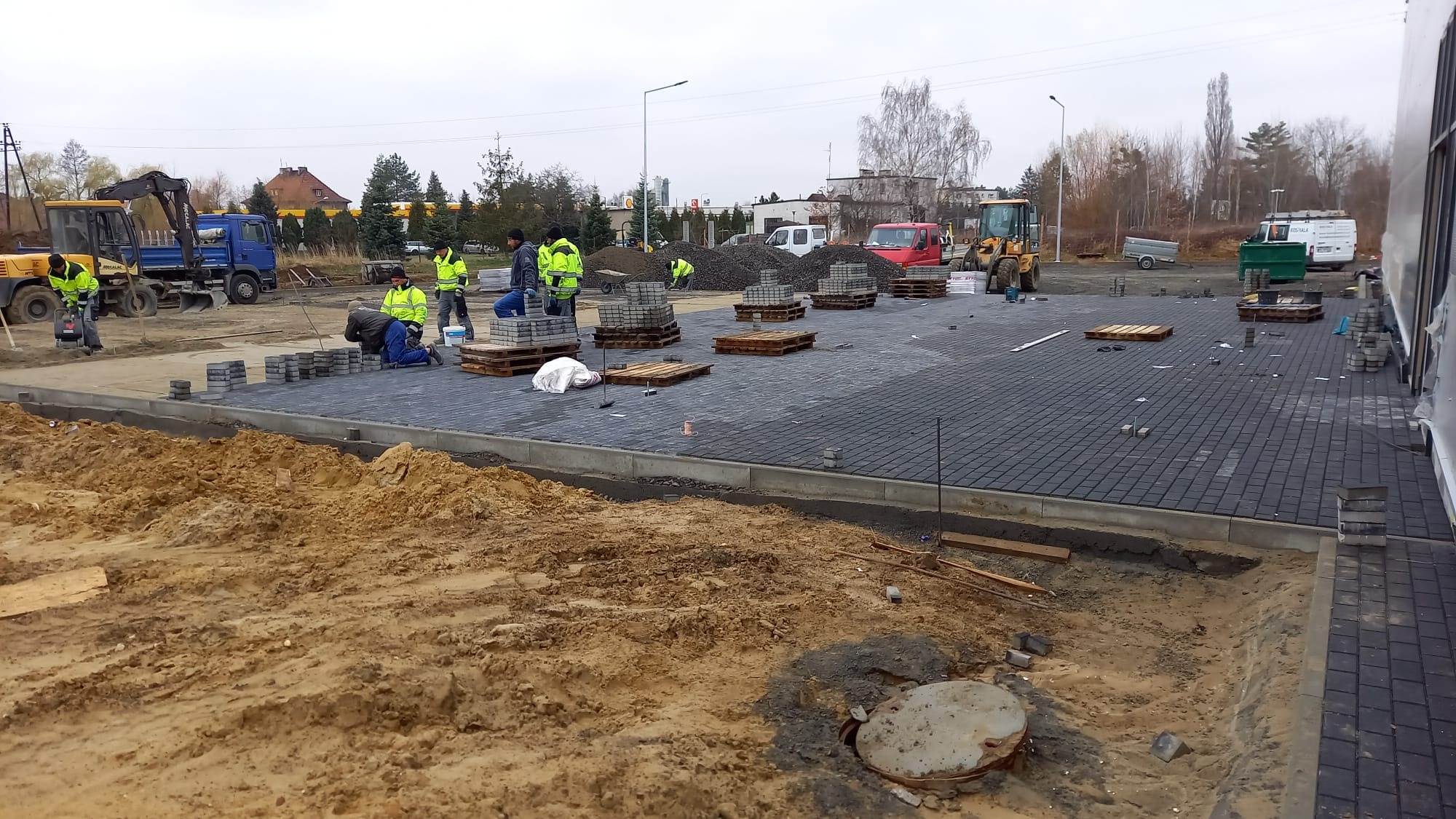 Układanie ciemnoszarej kostki brukowej na dużej powierzchni. Pracownicy w żółtych kamizelkach, maszyny budowlane w tle. Częściowo ułożona nawierzchnia.