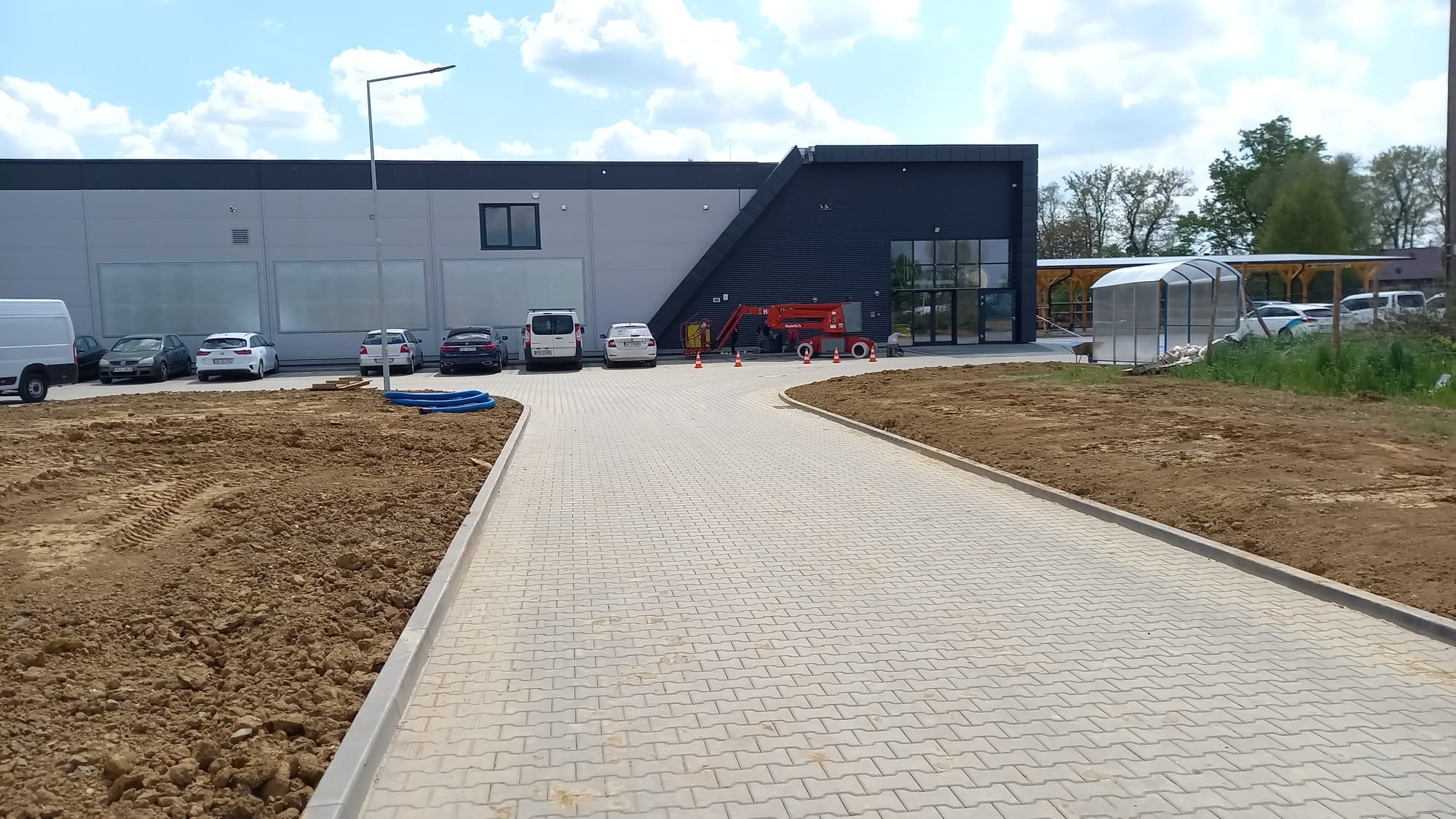 Nowa, jasnoszara kostka brukowa ułożona przed budynkiem komercyjnym. Widoczna maszyna budowlana i przygotowany teren pod dalsze prace. Słoneczny dzień.