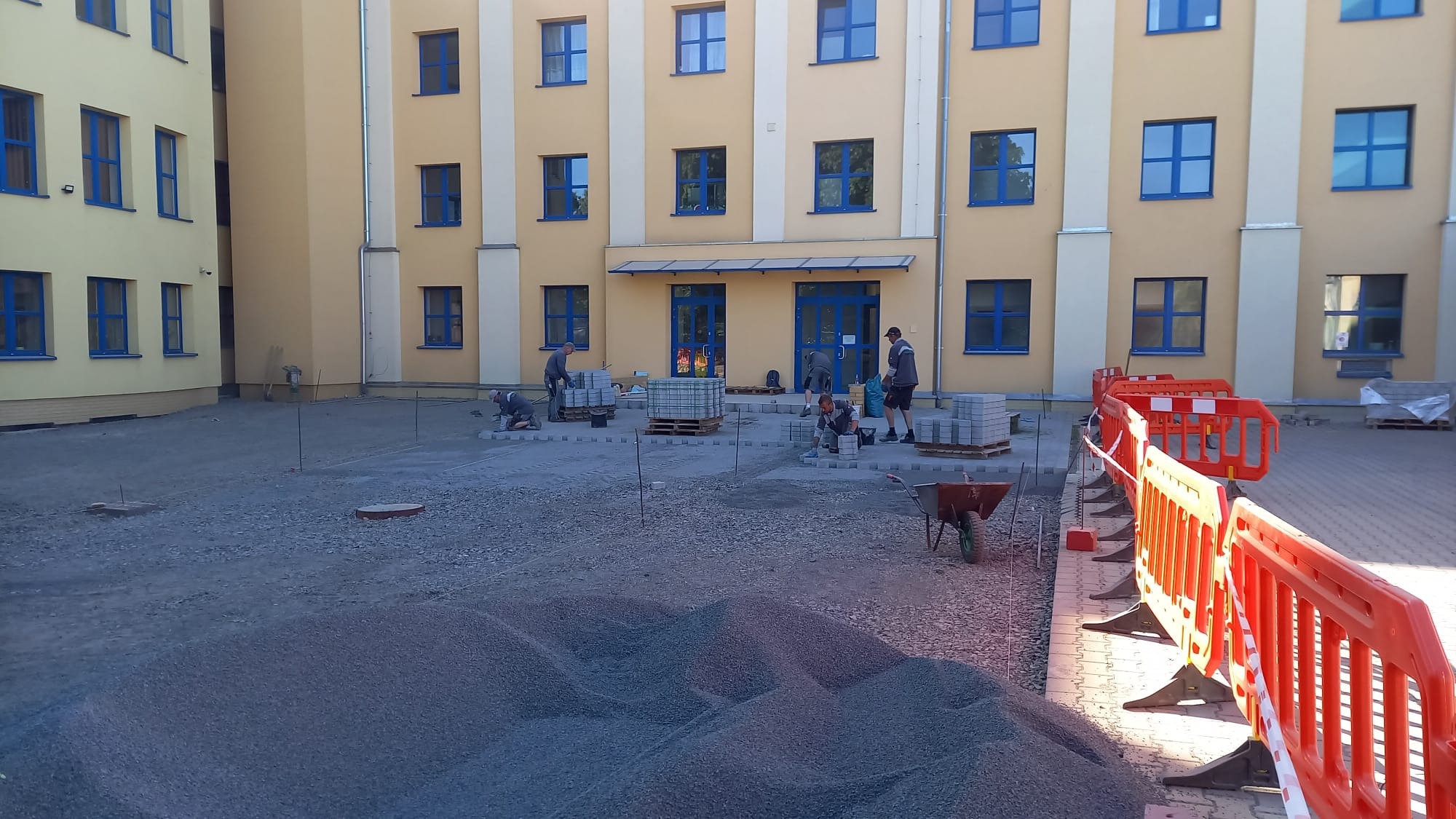 Praca wre: układanie kostki brukowej na placu przed budynkiem z niebieskimi oknami. Widoczne palety z kostką, narzędzia i robotnicy w trakcie pracy.