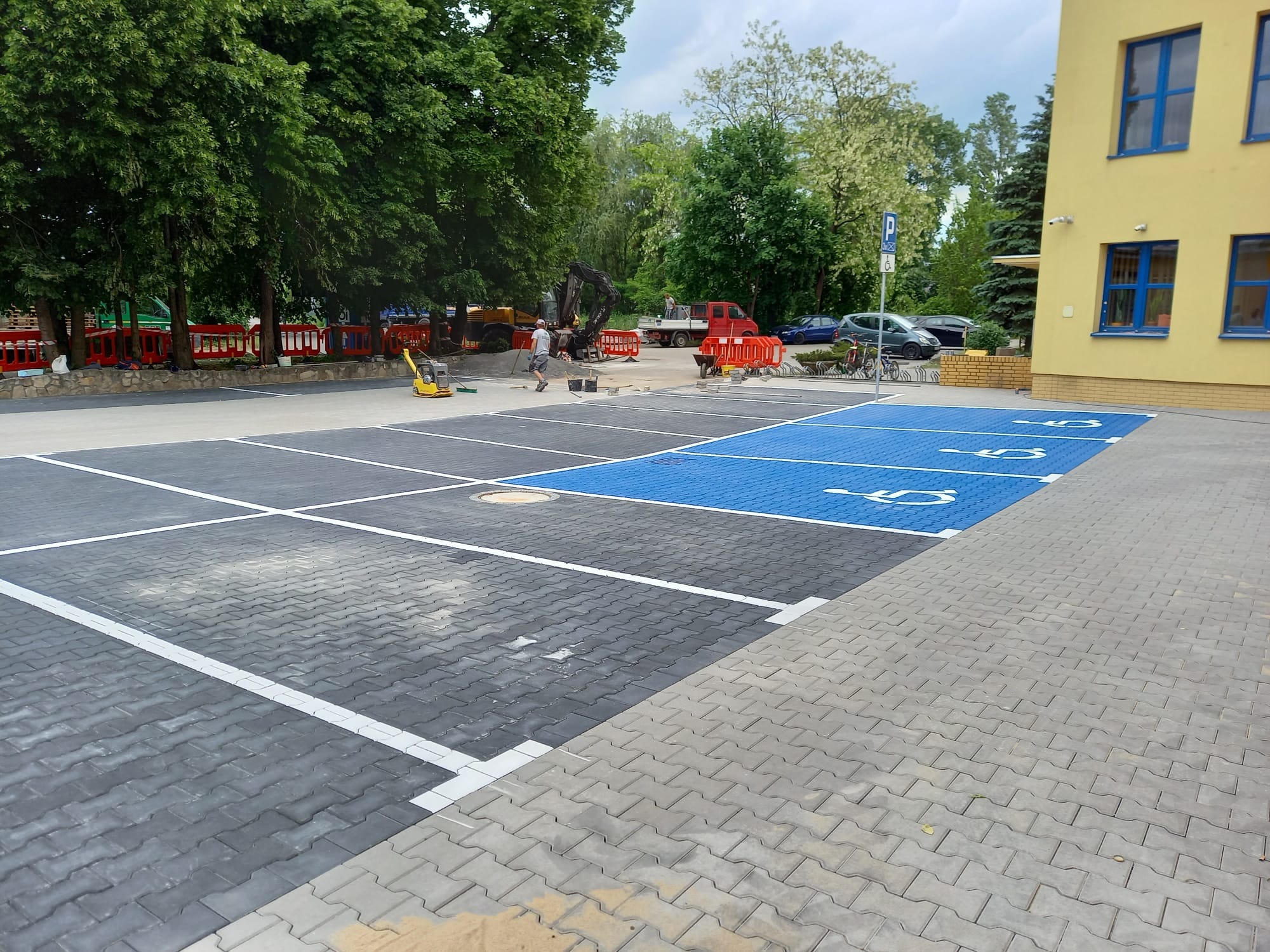Wykończony parking z kostki brukowej z wyznaczonymi miejscami dla niepełnosprawnych, w tle maszyna budowlana i pracownik. Część placu jeszcze nieukończona.