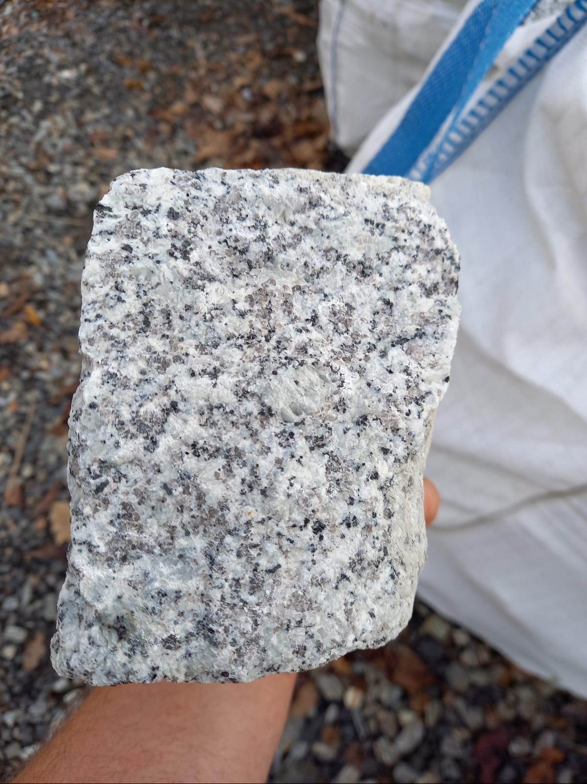Szary blok kostki granitowej o nieregularnych krawędziach, trzymany w dłoni, z widoczną strukturą kamienia i tłem z drobnym żwirem oraz workiem budowlanym.