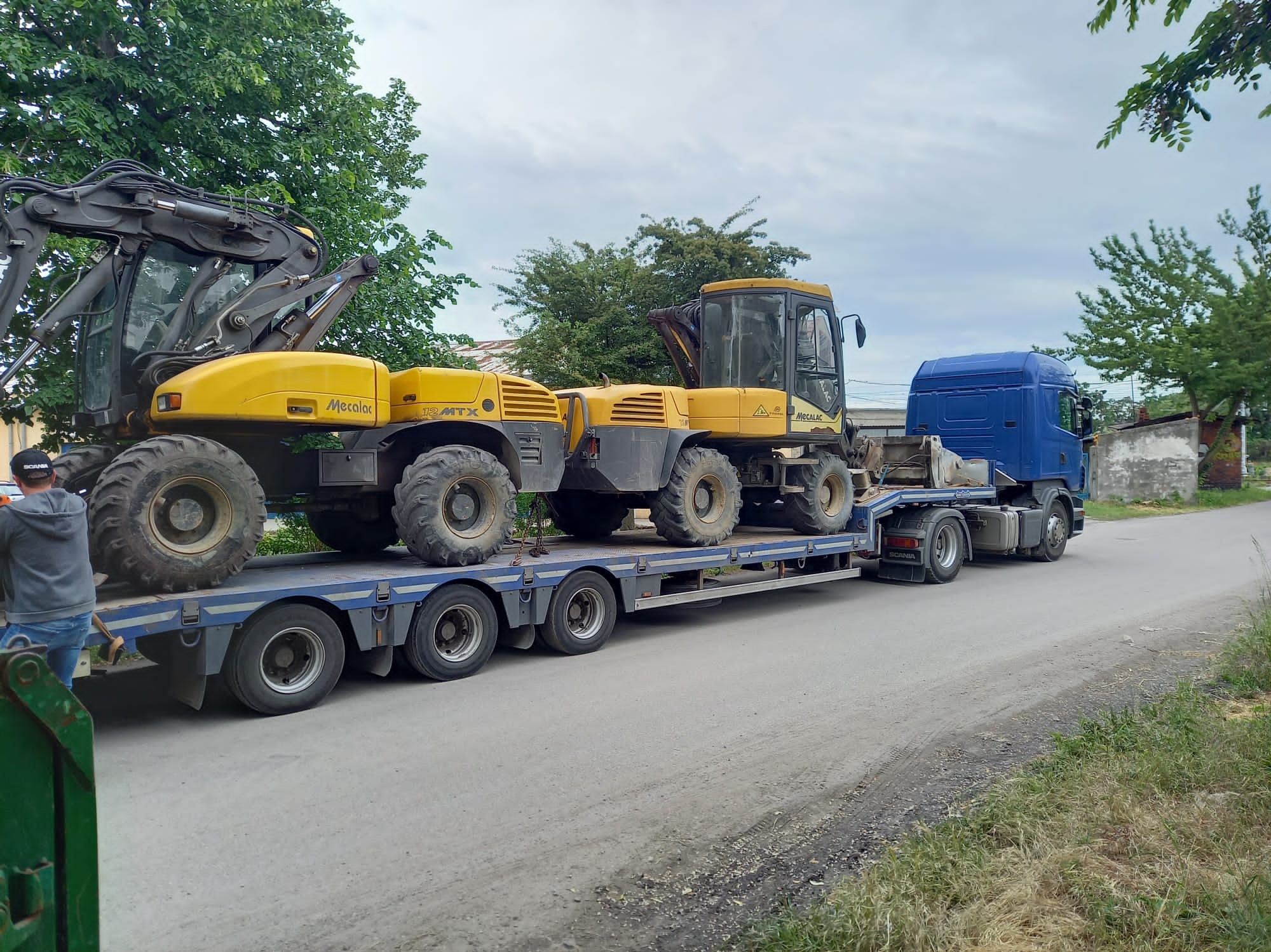Transport dwóch żółtych koparek Mecalac 12 MTX na niskopodwoziowej naczepie Scania. Widok z boku na asfaltową drogę w słoneczny dzień.
