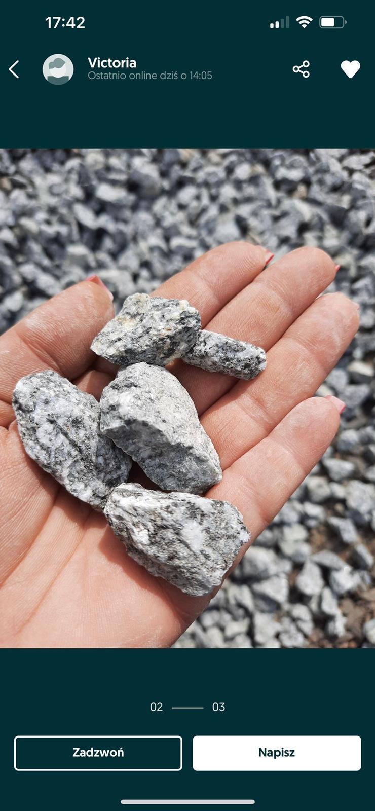Dłoń trzymająca kilka bryłek szarej kostki granitowej na tle wysypanego żwiru. Widoczne drobne użylenia i struktura kamienia.