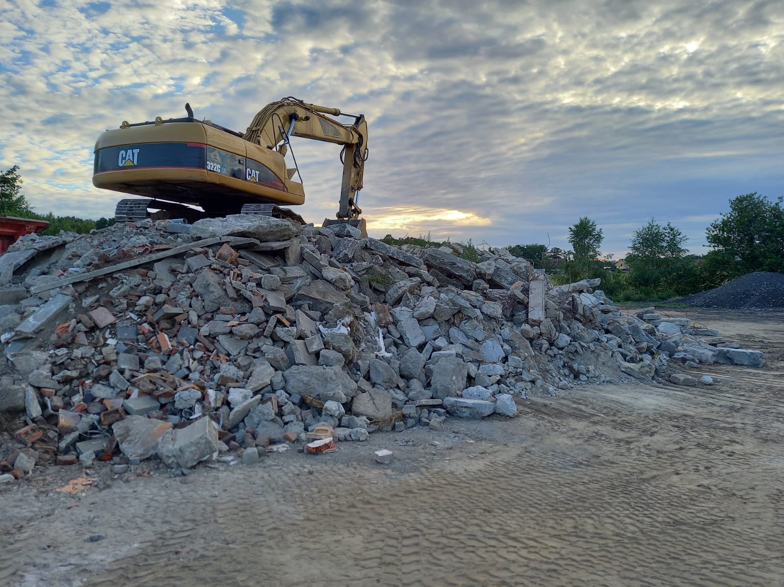 Żółty koparka CAT 322C LR na stercie gruzu i cegieł pod pochmurnym niebem. Widoczne ślady opon na ziemi. Prace rozbiórkowe w toku.