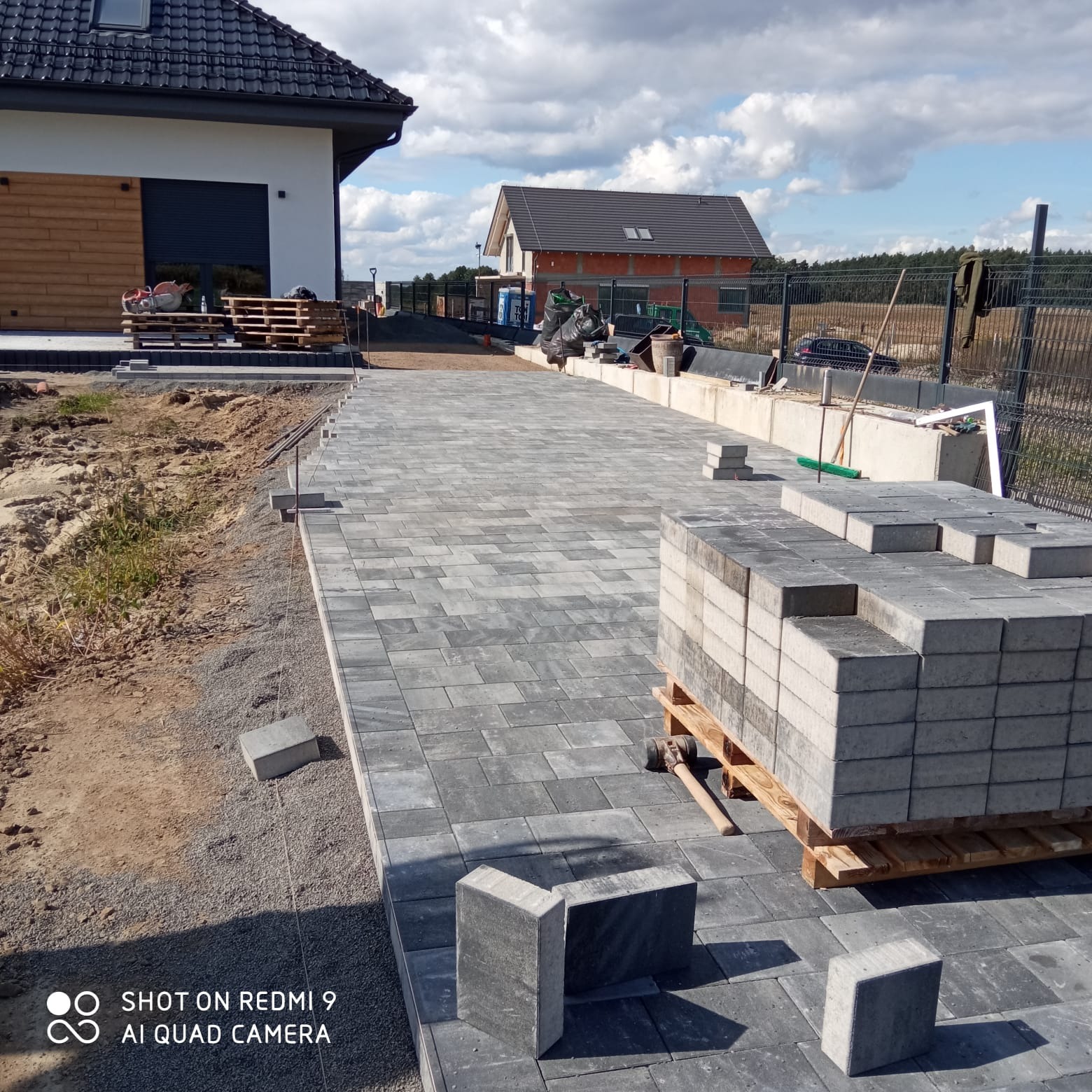 Układanie szarej kostki brukowej na podjeździe w Ligocie Prószkowskiej. Częściowo wyłożona nawierzchnia, paleta kostki brukowej, młotek brukarski. Widok na domy i ogrodzenie.
