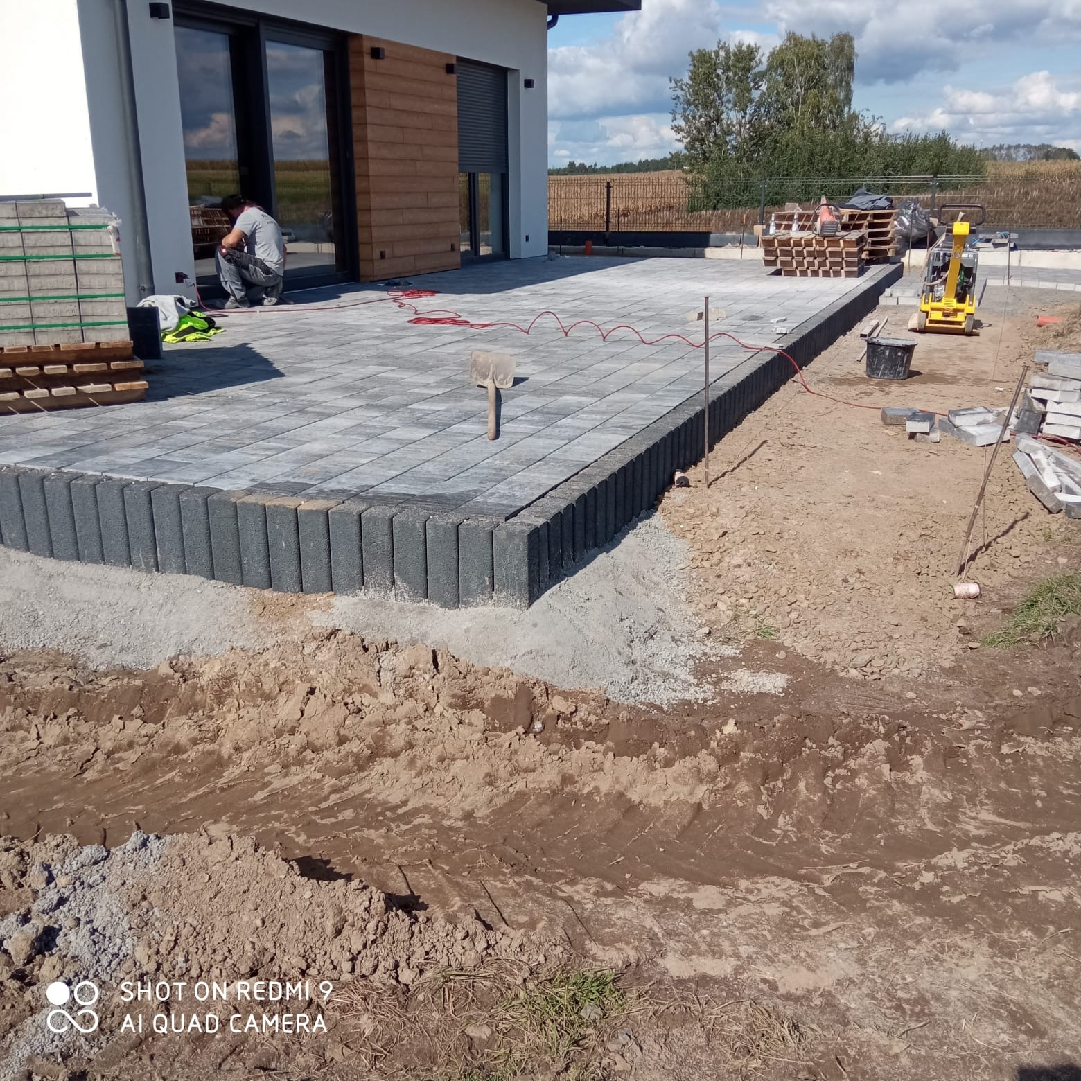 Częściowo ułożona, szara kostka brukowa z obrzeżami na posesji. W tle dom z dużymi oknami, palety z kostką i zagęszczarka. Widoczny robotnik w trakcie pracy.