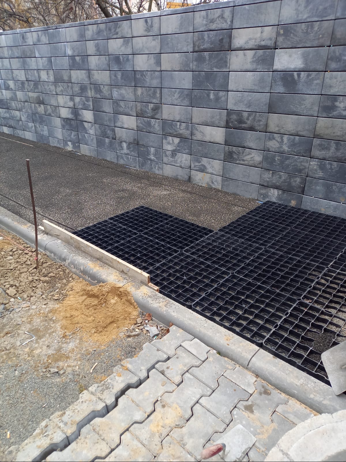 Etap prac: układanie kratki trawnikowej i kostki przy betonowym obrzeżu, z murem oporowym z szarej kostki w tle. Widoczne materiały budowlane.