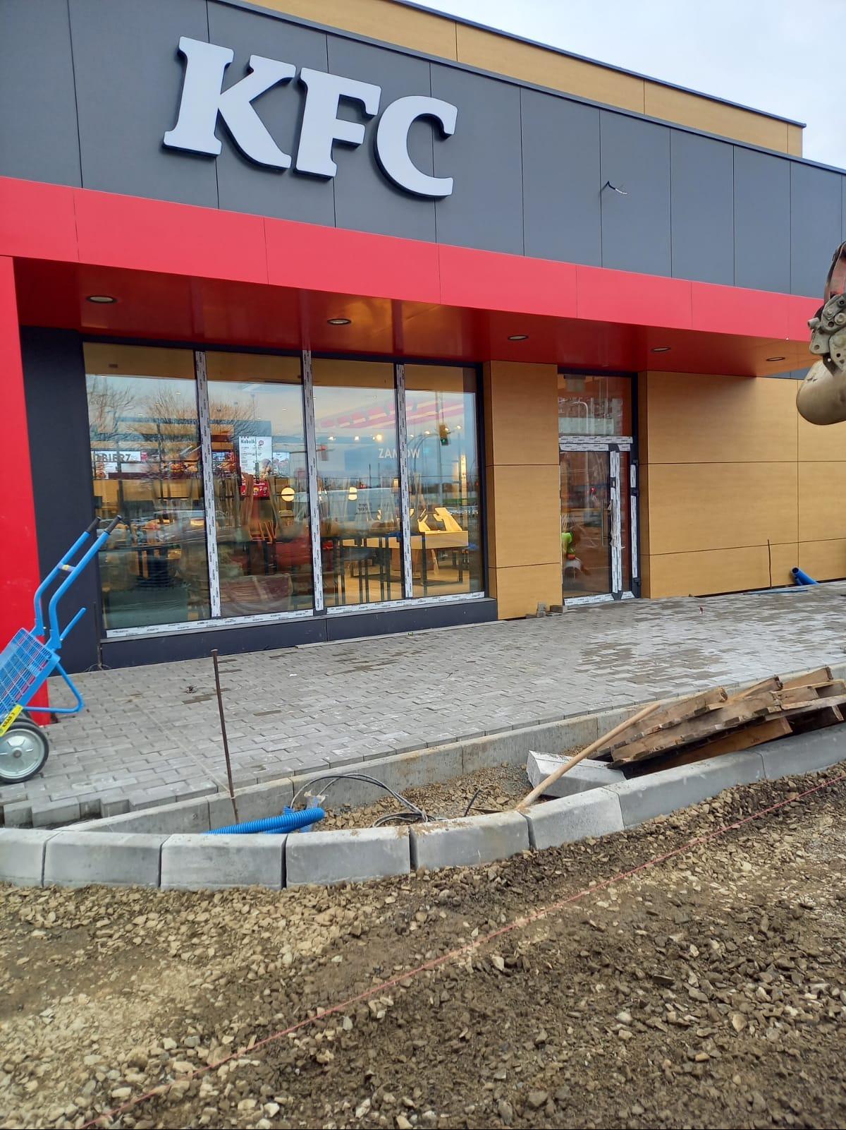 Układanie obrzeży i kostki brukowej przed budynkiem KFC. Widoczne materiały budowlane, narzędzia i fragment koparki. Etap prac brukarskich.