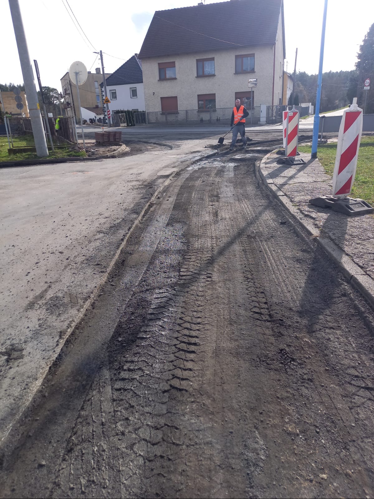Pracownik w kamizelce odblaskowej wyrównuje łopatą teren pod drogę. Widoczne ślady opon i rozłożone materiały. W tle budynki mieszkalne i słup.