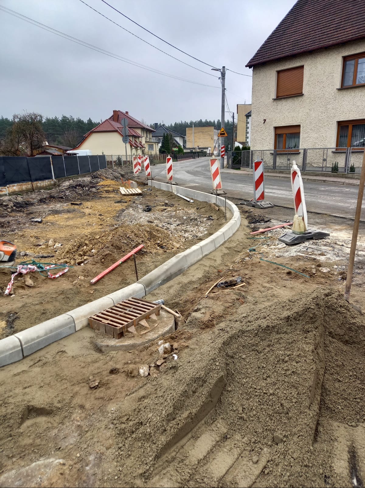 Montaż obrzeży betonowych na drodze, widoczne narzędzia, hałdy piasku i zabezpieczenia drogowe. Etap przygotowania podłoża pod kostkę.