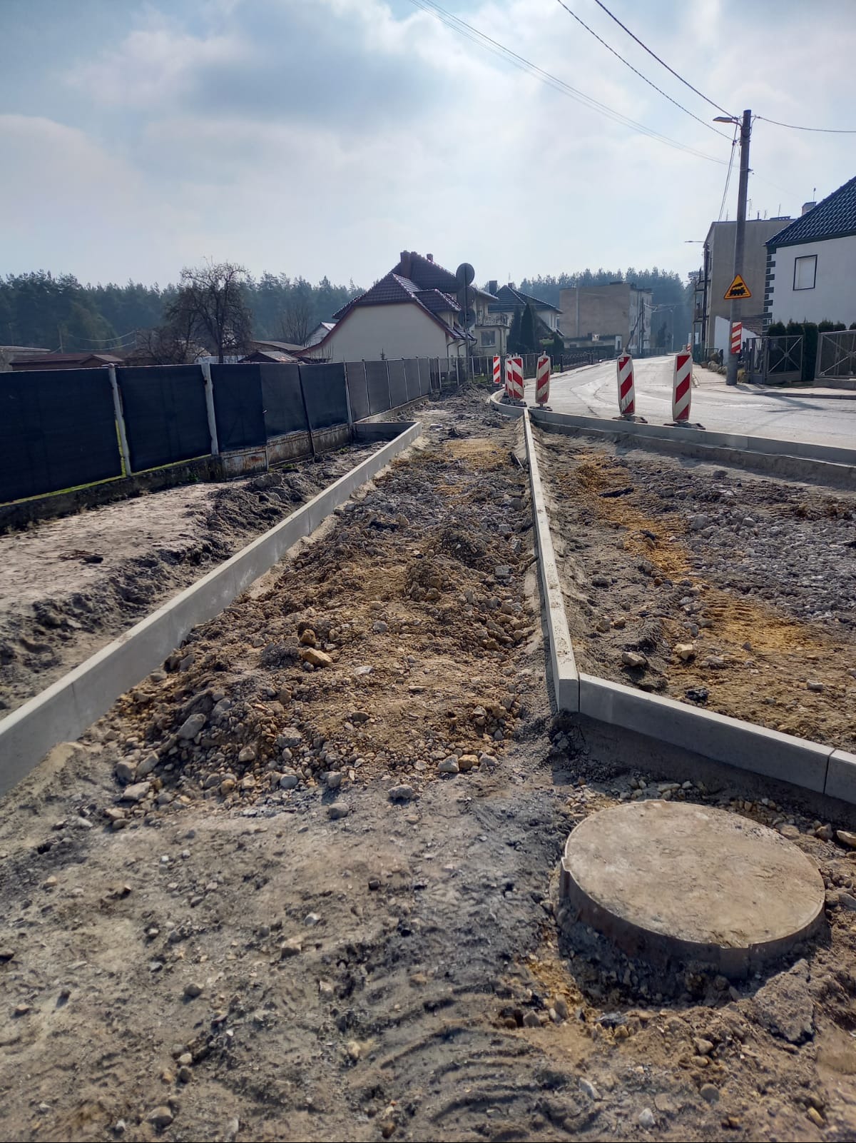 Przygotowanie podłoża pod chodnik z obrzeżami na tle zabudowy i drzew. Widoczne znaki drogowe.