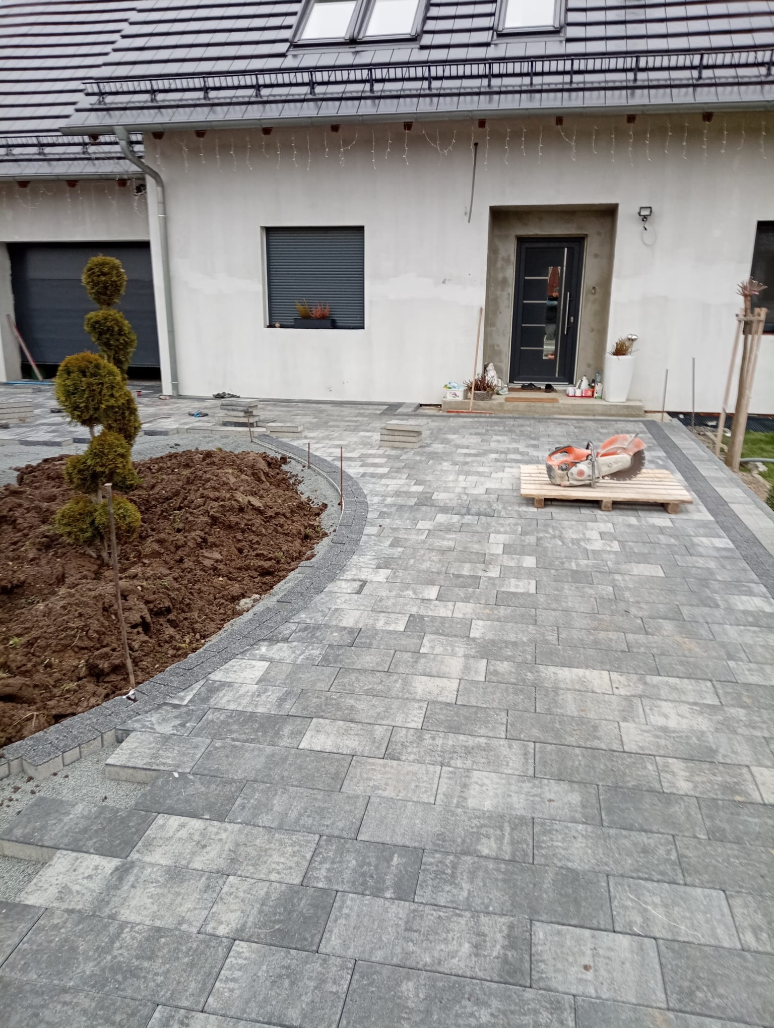 Ułożona szara kostka brukowa przed domem z narzędziem do cięcia na palecie. W tle widoczny fragment budynku z oknem i drzwiami wejściowymi.