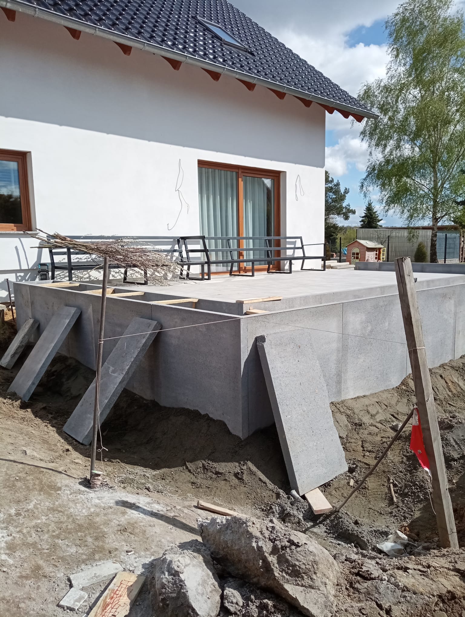Betonowy taras w trakcie budowy przy białym domu z ciemnym dachem. Widoczne podpory i przygotowany teren. W tle drzewo i błękitne niebo.