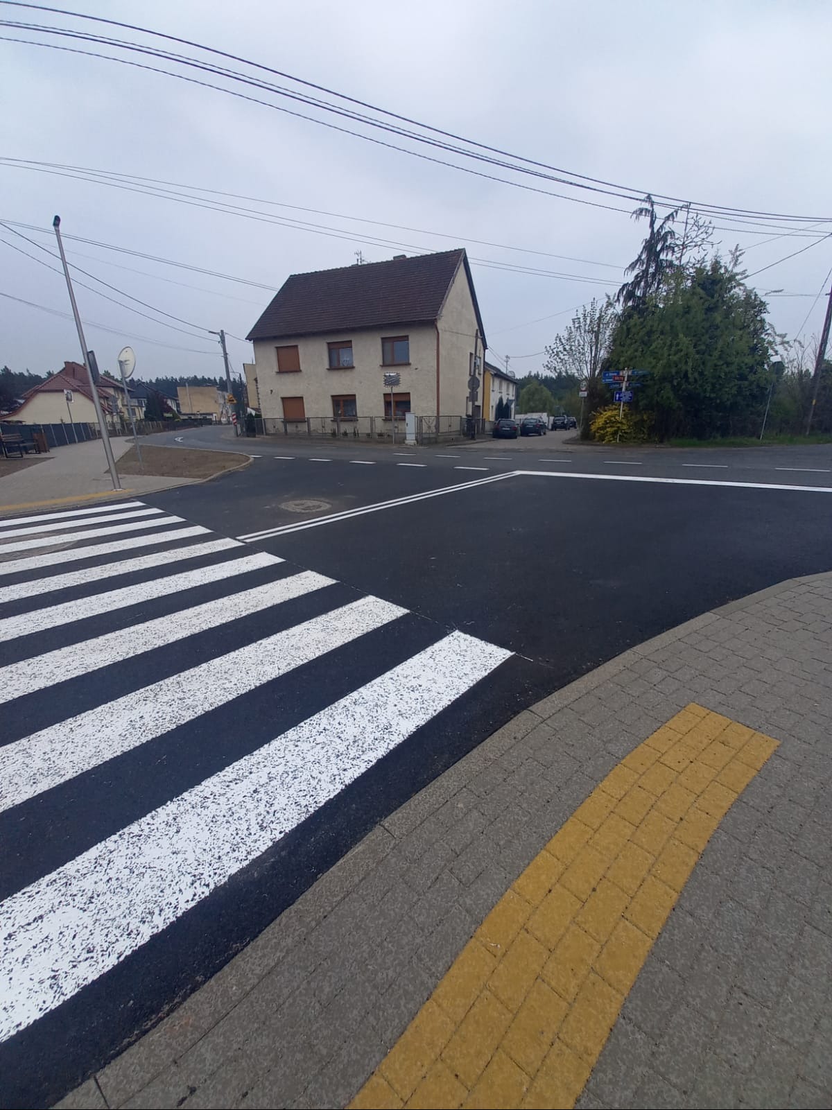 Nowe przejście dla pieszych z żółtymi płytami ostrzegawczymi na chodniku, świeżo pomalowane pasy zebry na tle wiejskiego krajobrazu.