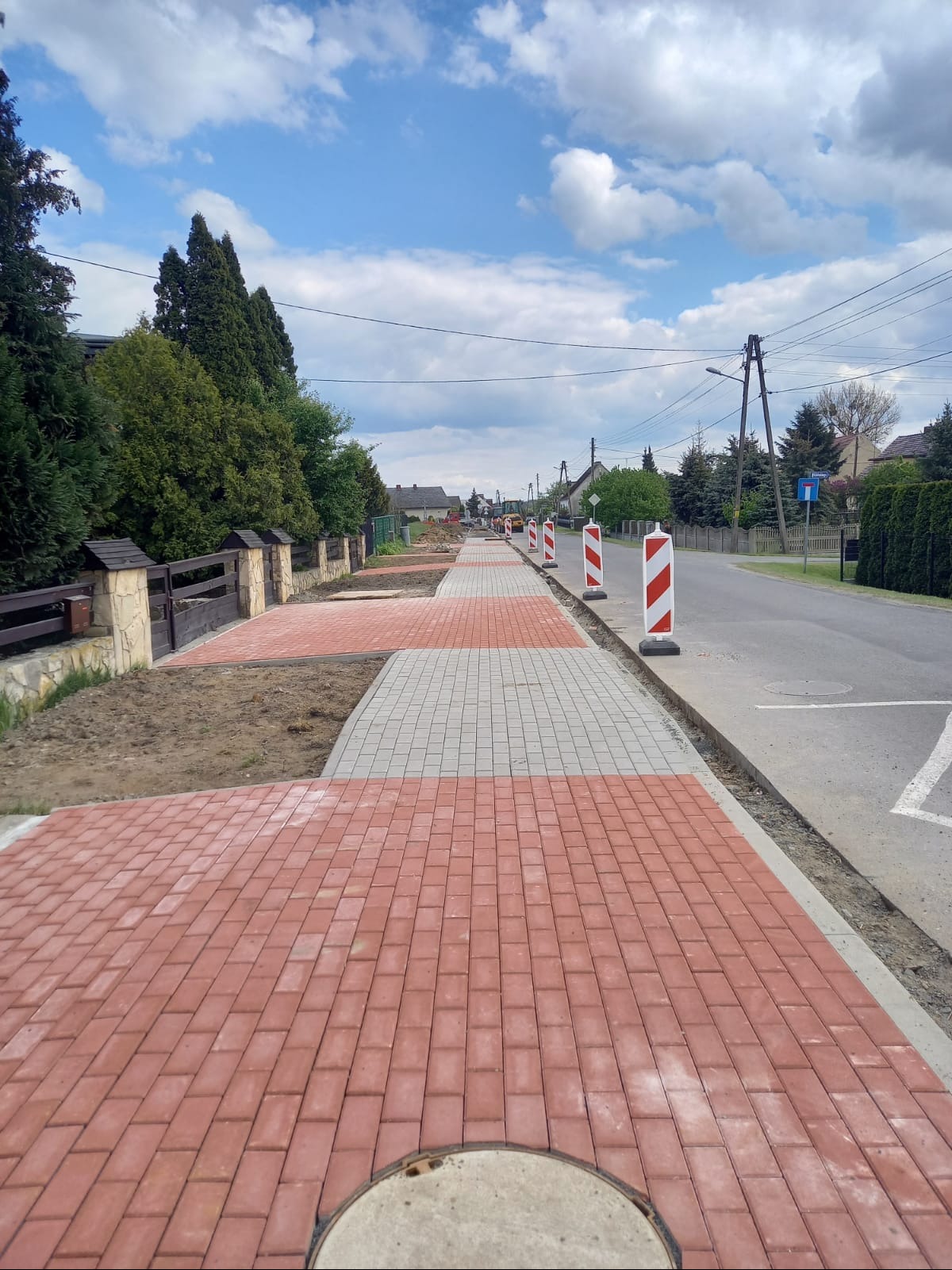 Częściowo ułożony chodnik z czerwonej i szarej kostki brukowej wzdłuż ulicy, zabezpieczony słupkami ostrzegawczymi. Prace brukarskie w toku, widoczne przygotowane podłoże.