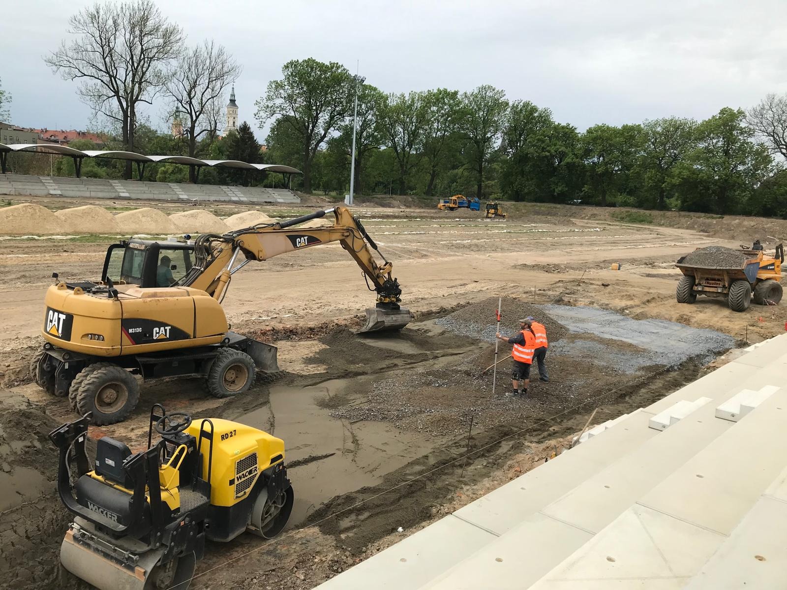 Żółty walec Wacker i koparka CAT na placu budowy stadionu, gdzie robotnicy w kamizelkach mierzą teren. W tle trybuny i inne maszyny.