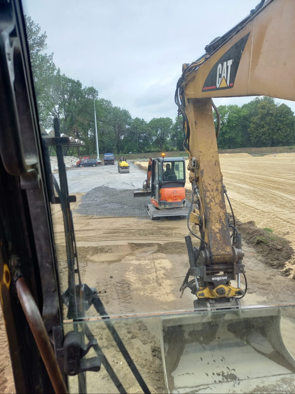 Widok z kabiny na prace ziemne: mini koparka Kubota wysypuje żwir, walec drogowy ubija teren. W tle zaparkowany samochód ciężarowy i przygotowany teren pod budowę.