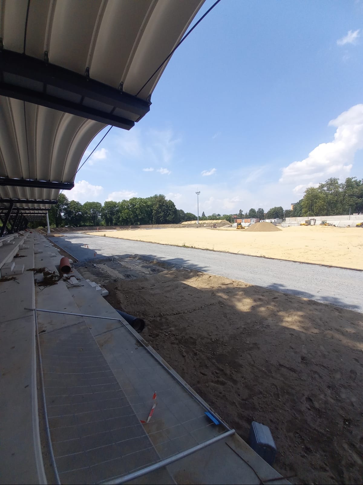 Widok na plac budowy z trybun stadionu: przygotowany teren pod nawierzchnię, maszyny budowlane w tle, częściowo zadaszone trybuny.