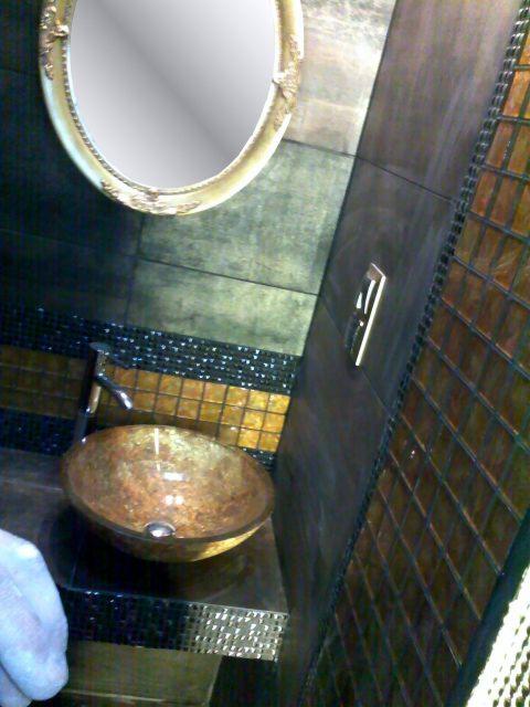 Wykończona łazienka z umywalką w odcieniach brązu, zdobionym lustrem i mozaiką. Ciemne płytki na ścianach i blacie, detale w kolorze złota i srebra.