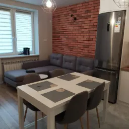 Jasne wnętrze mieszkania z ceglaną ścianą, szarą sofą, białym stołem jadalnym i nowoczesnymi lampami wiszącymi, prezentujące efekt malowania i wykończenia.