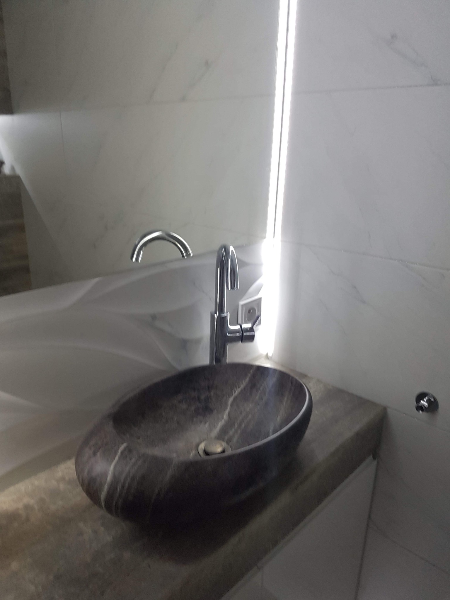 Owalna umywalka z ciemnego kamienia z chromowaną baterią na blacie z jasnego kamienia, podświetlona listwą LED w łazience wykończonej marmurowymi płytkami.