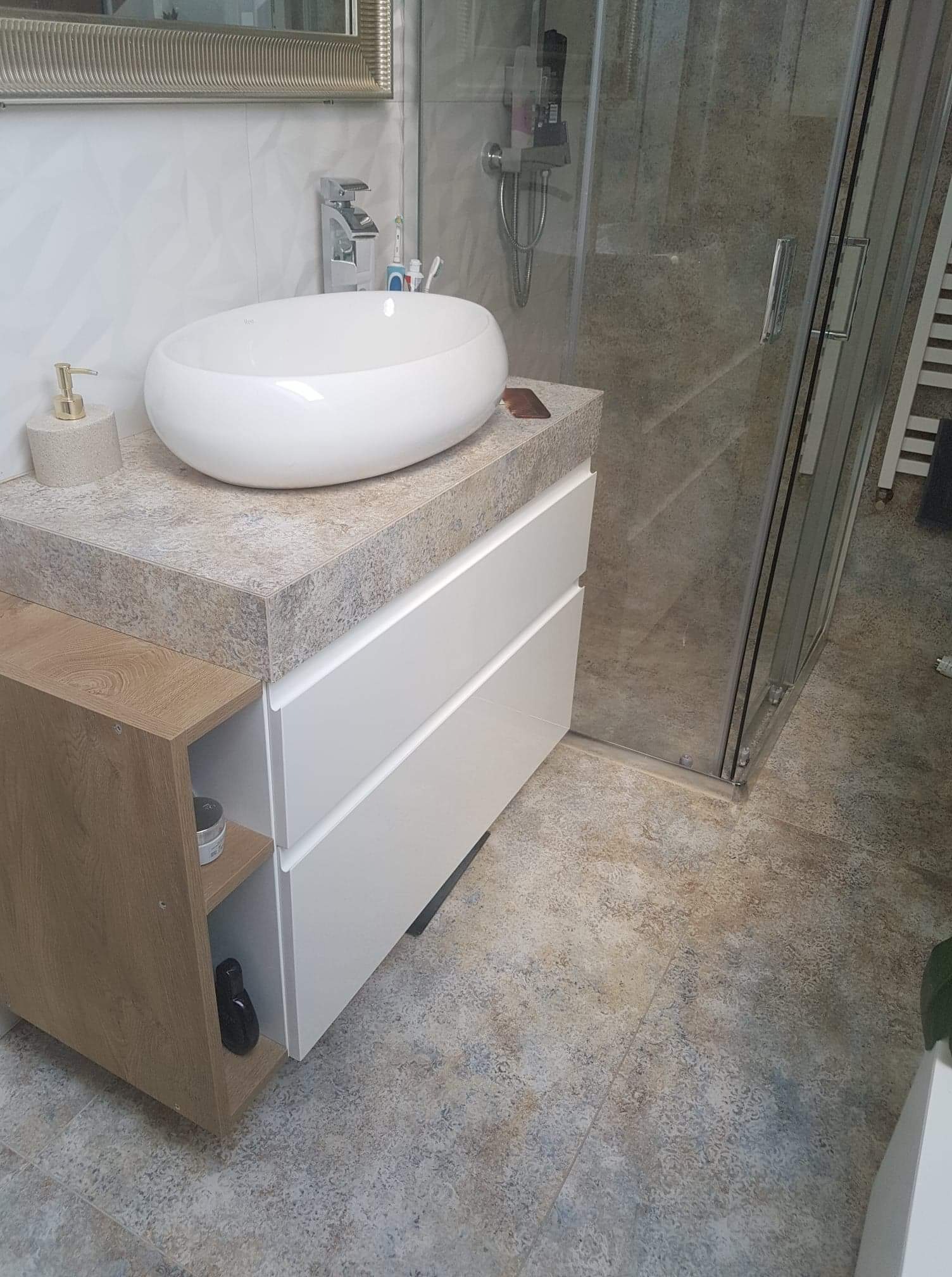 Nowoczesna łazienka z owalną umywalką nablatową, białą szafką z szufladami i kabiną prysznicową z szarymi płytkami.