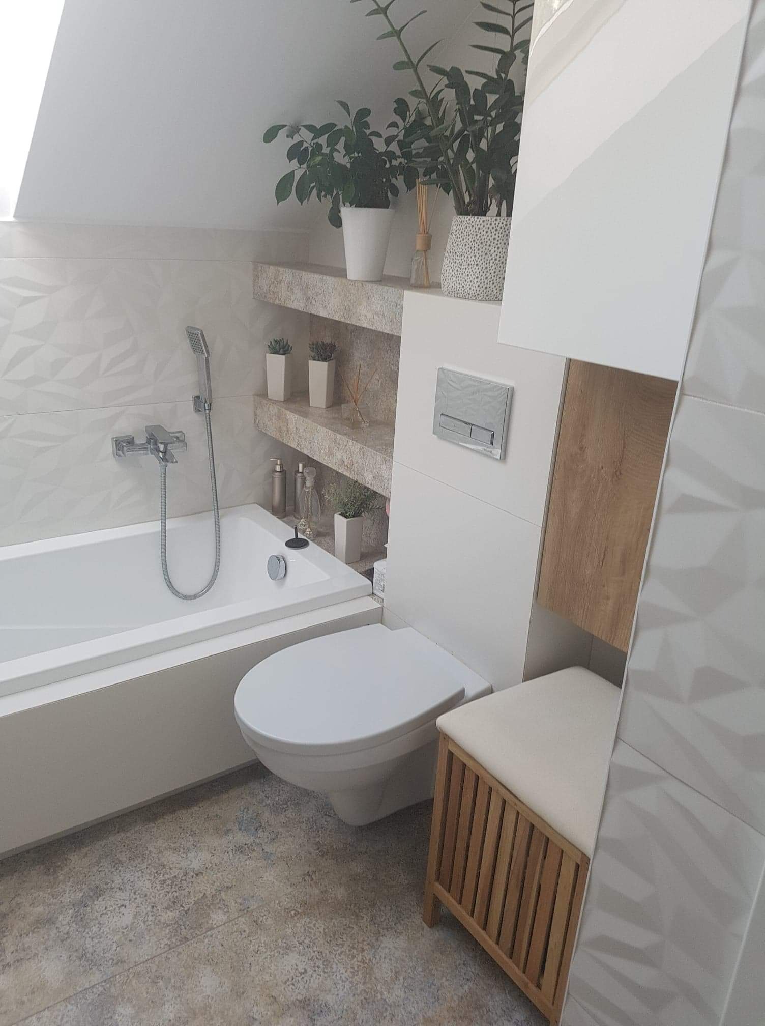 Jasna łazienka na poddaszu z wanną, toaletą, drewnianym siedziskiem i półkami w ścianie z dekoracjami roślinnymi.