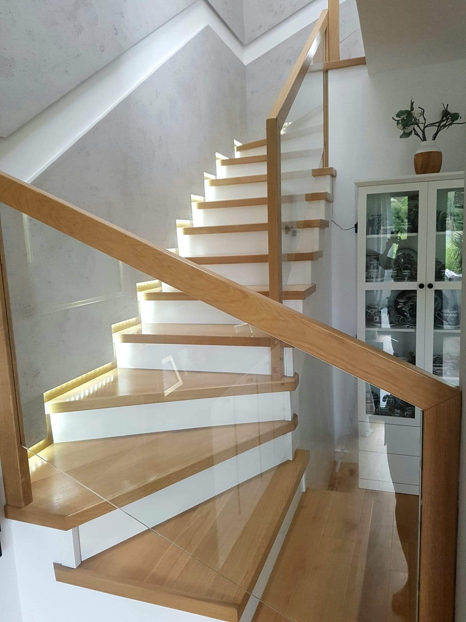 Nowoczesne schody drewniane z białymi podstopnicami i szklaną balustradą, podświetlane od spodu, widok z dołu na tle ściany o fakturze betonu i białej szafki z ekspozycją.