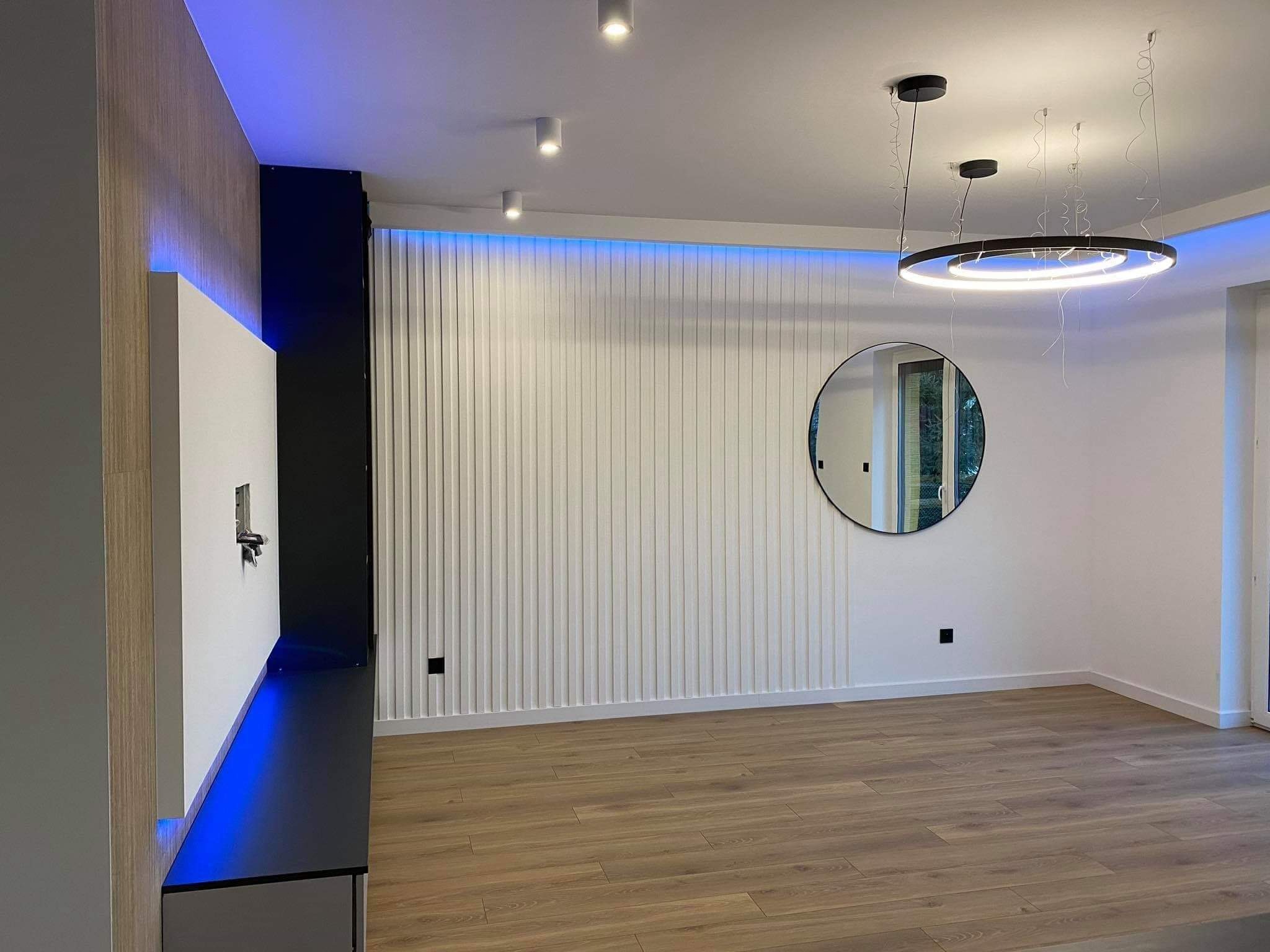 Nowoczesne wnętrze z drewnianą podłogą, ścianą z pionowymi panelami i okrągłym lustrem, oświetlone listwą LED i okrągłą lampą wiszącą.