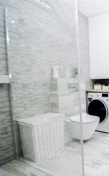 Jasna łazienka z szarymi płytkami imitującymi kamień, białą toaletą wiszącą, koszem wiklinowym i pralką.