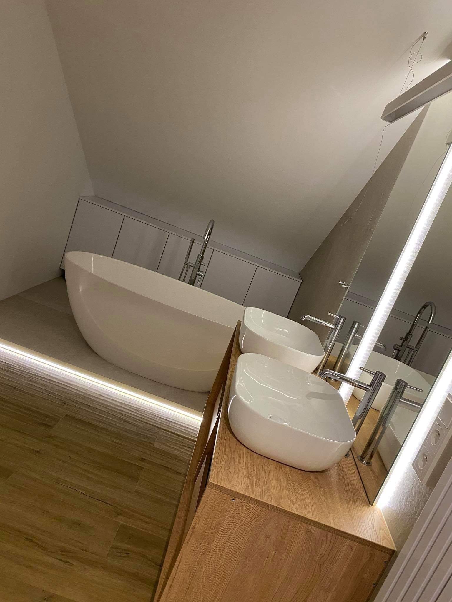 Nowoczesna łazienka na poddaszu z dwiema owalnymi umywalkami nablatowymi, wolnostojącą wanną i podświetleniem LED pod wanną.