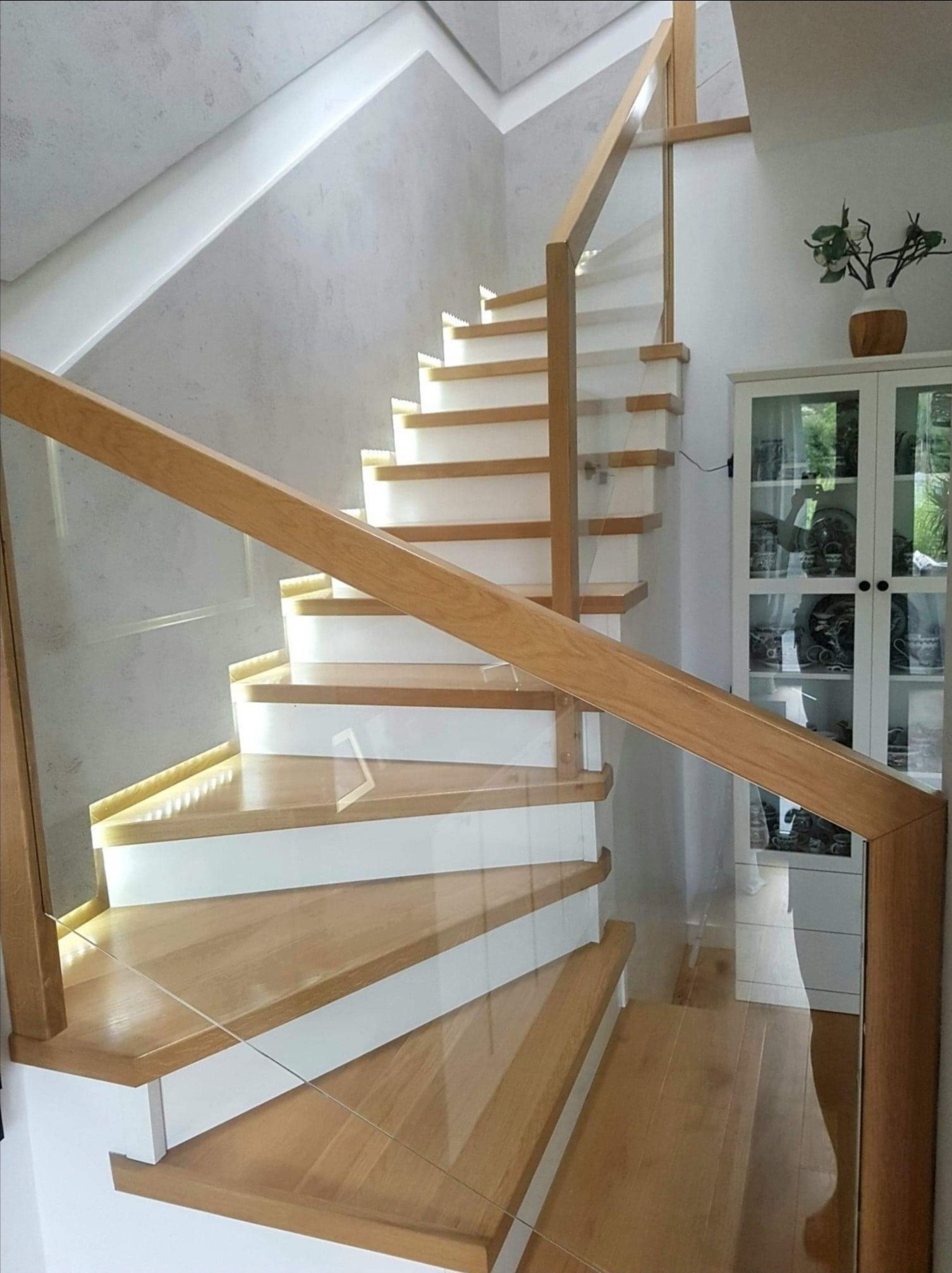 Nowoczesne schody drewniane z białymi podstopnicami i szklaną balustradą, oświetlenie LED pod stopniami, widok na białą witrynę z dekoracjami.