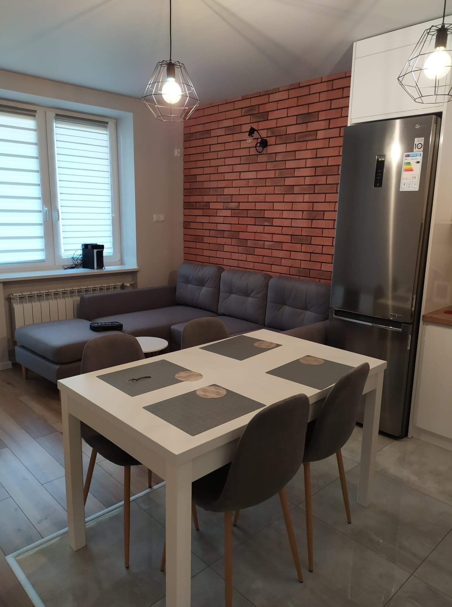 Jasne wnętrze mieszkania z ceglaną ścianą, szarą sofą, białym stołem jadalnym i nowoczesnymi lampami wiszącymi, prezentujące efekt malowania i wykończenia.