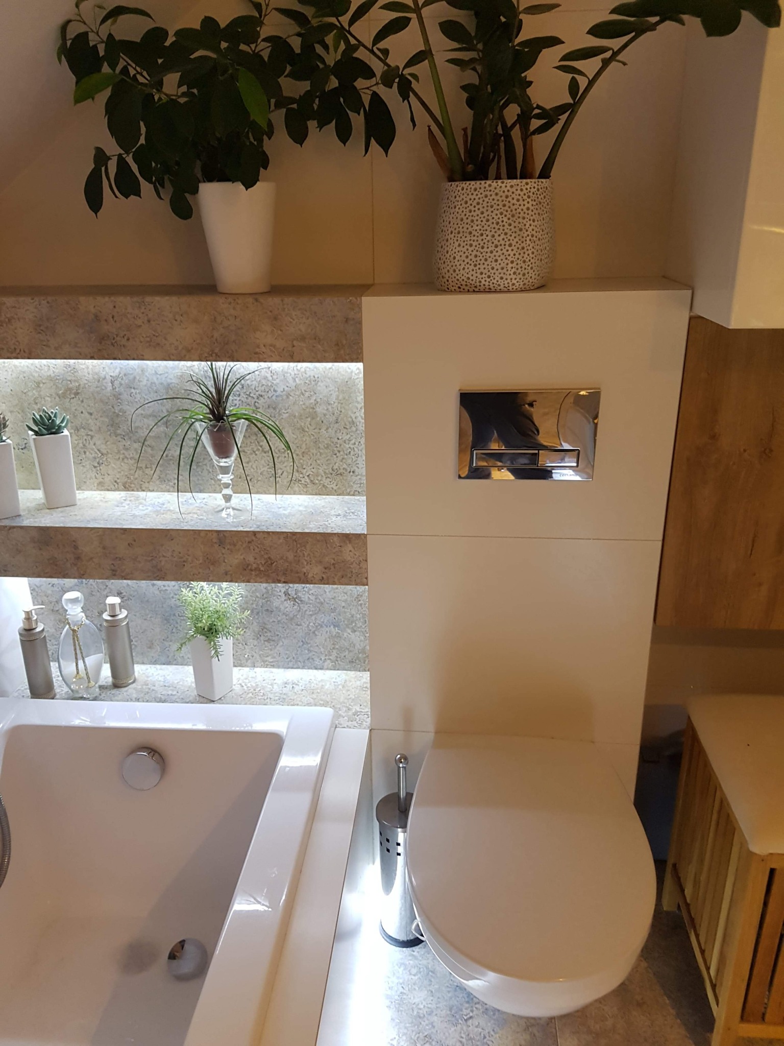 Nowoczesna łazienka z podświetlanymi półkami, roślinami doniczkowymi, białą wanną i toaletą podwieszaną z chromowanym przyciskiem spłukującym.