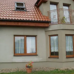 Dom z oknami PCV w drewnopodobnej okleinie i balkonem z kutą balustradą, widok z zewnątrz, z kwiatami w doniczce na pierwszym planie.