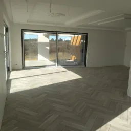 Jasne, przestronne wnętrze z ułożoną w jodełkę podłogą z płytek ceramicznych imitujących drewno, duże przesuwne okna z widokiem na zewnątrz, sufit z ozdobnymi sztukateriami i widocznymi przewodami...