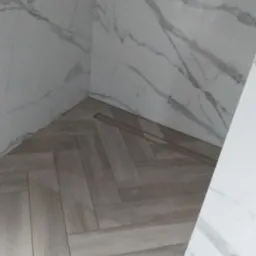 Jasne, drewnopodobne płytki ułożone w jodełkę na podłodze, z odpływem liniowym. Ściany wyłożone białymi płytkami imitującymi marmur.