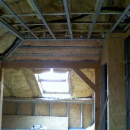 Poddasze w trakcie prac: izolacja wełną mineralną, konstrukcja z płyt OSB i metalowych profili, widoczne okno dachowe.