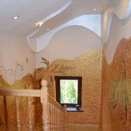 Klatka schodowa we Władysławowie z oryginalną dekoracją ścian imitującą krajobraz, drewniane schody i balustrada, sufit z podwieszanymi elementami i oświetleniem punktowym.