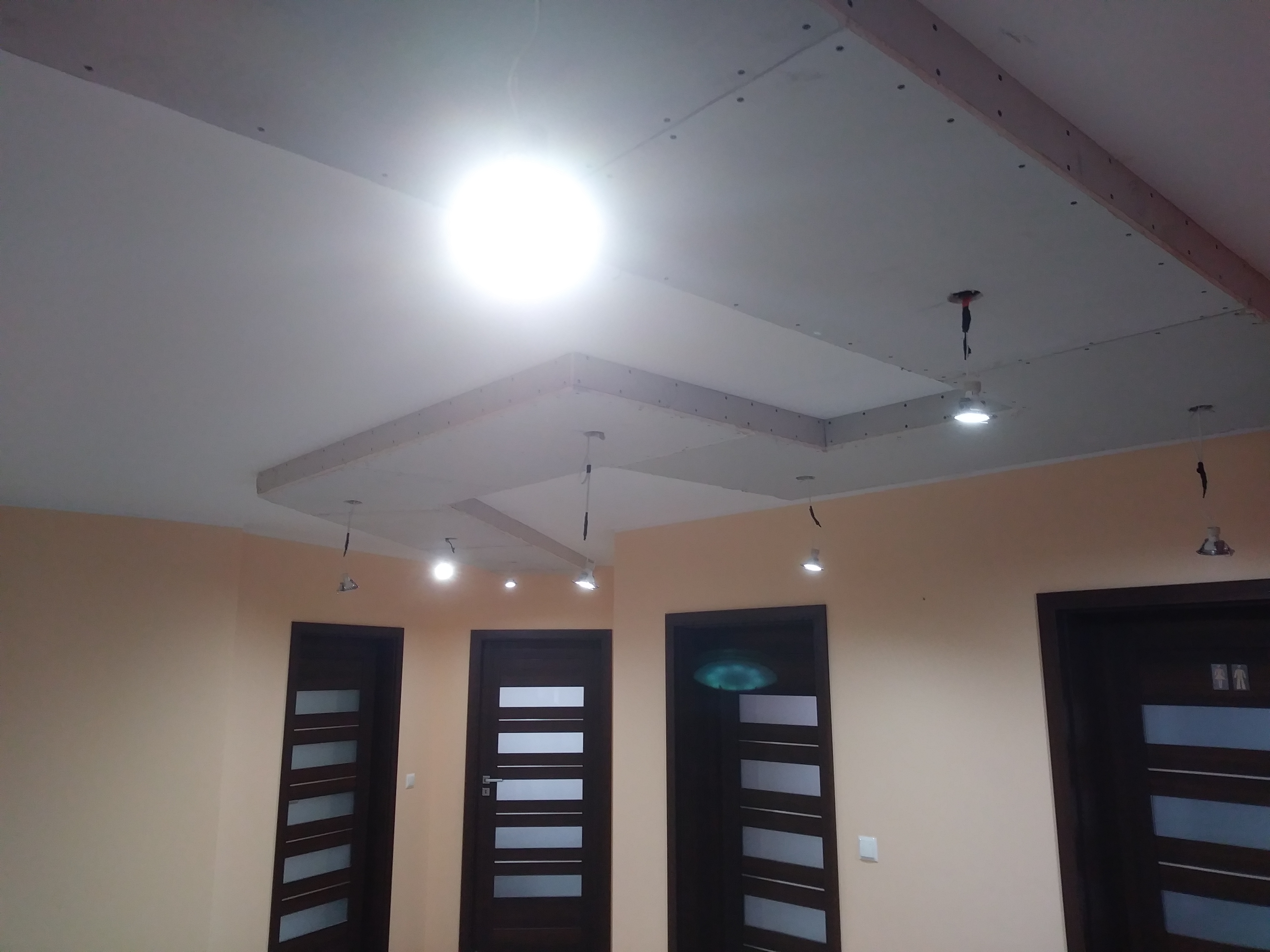 Sufit z płyt kartonowo-gipsowych w trakcie montażu, widoczne profile i otwory na oświetlenie, ściany w kolorze beżowym, ciemne drzwi z przeszkleniami.