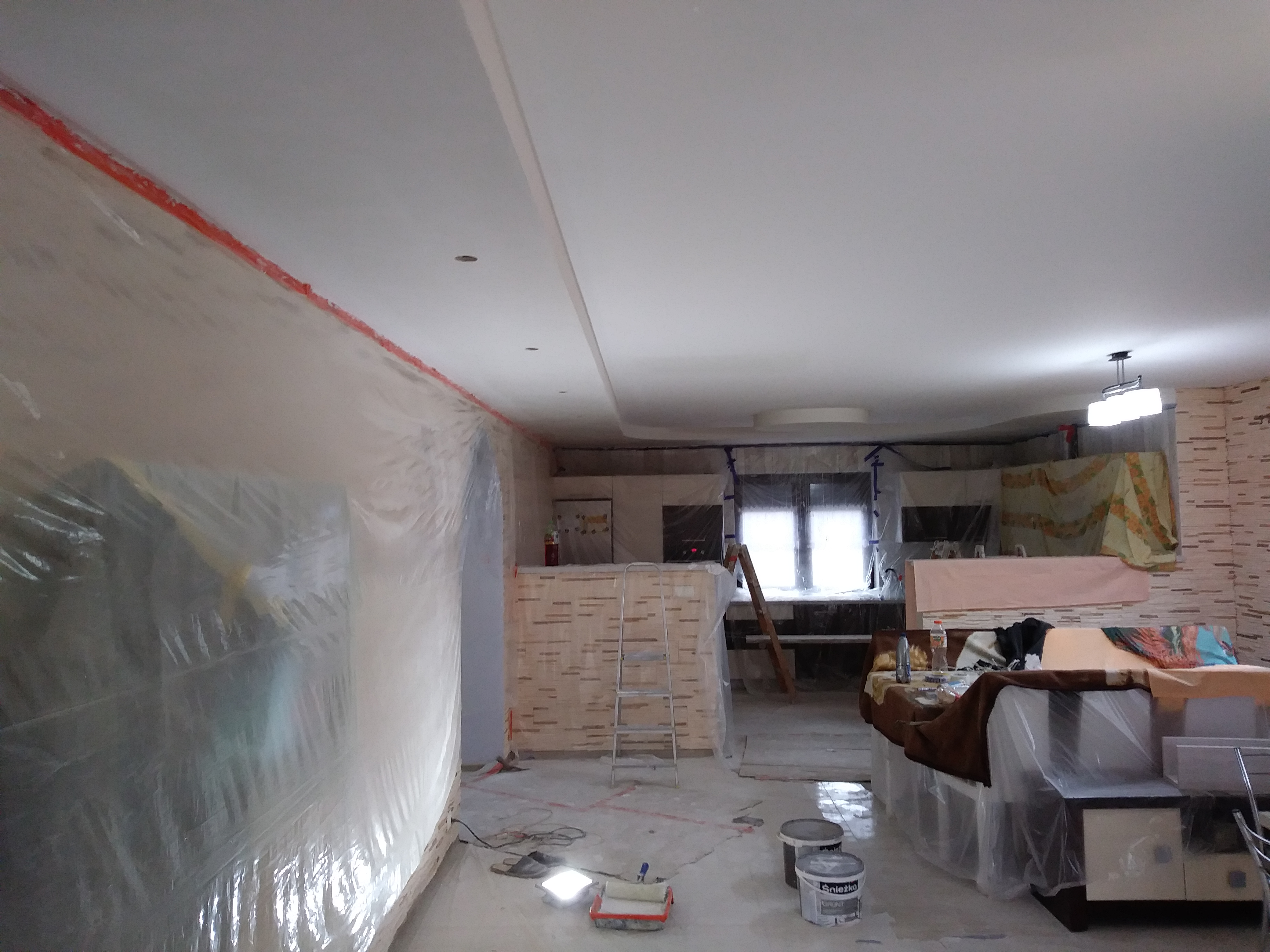 Pokój w trakcie zabezpieczania folią malarską, widoczne narzędzia malarskie i drabina, przygotowania do malowania ścian.
