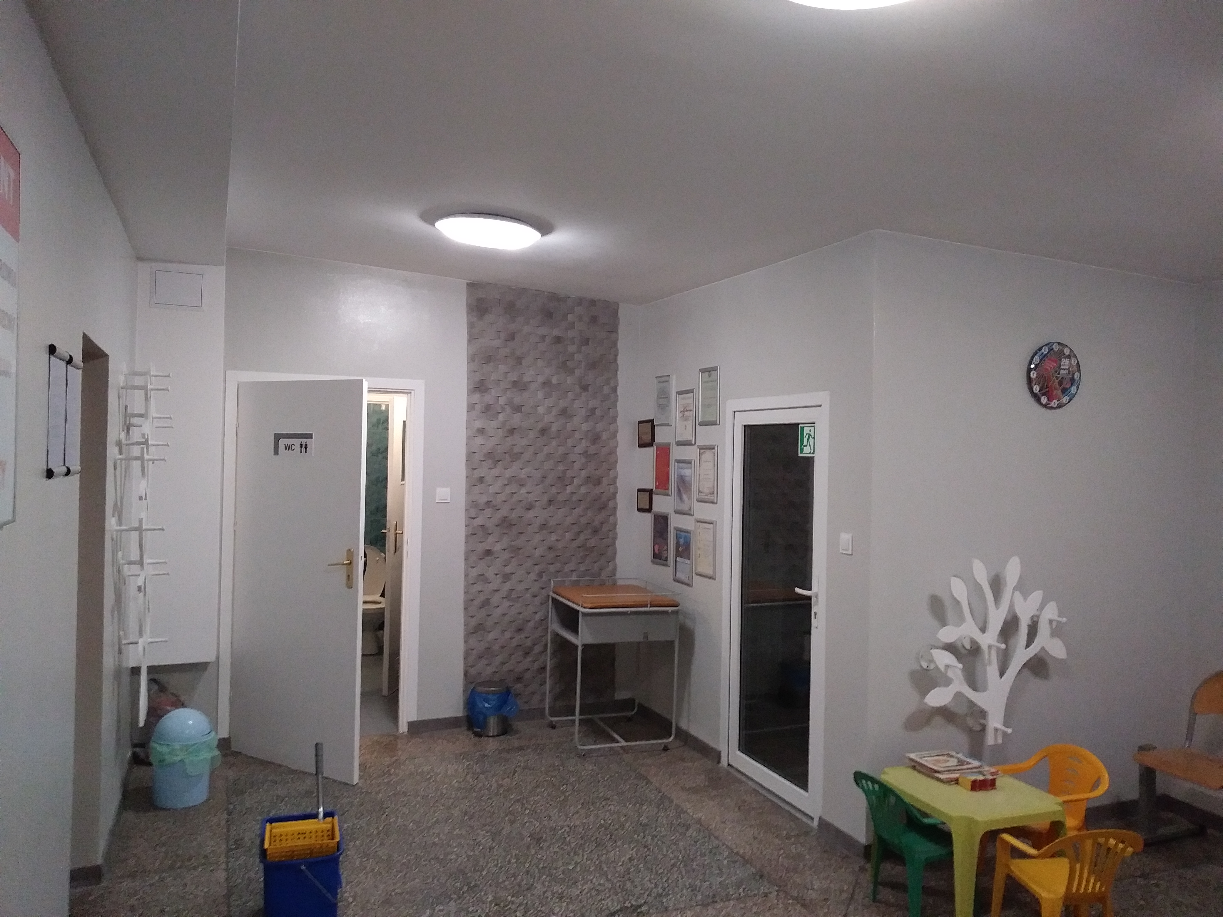 Jasne wnętrze z szarą mozaiką na ścianie, widoczne drzwi do toalety, stolik dla dzieci z krzesłami i dekoracyjny wieszak w kształcie drzewa.