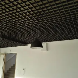 Czarne, kratowe wykończenie sufitu podwieszanego w jasnym pomieszczeniu z białymi ścianami, wiszące lampy w kształcie kropli i widoczne wejście ze schodami.