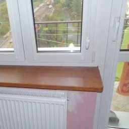 Olaf Wiewióra AWO SERWIS - Wykończony parapet z drewna pod białym oknem PCV. Widok na zewnątrz z suszącym się praniem. Grzejnik pod parapetem. Fragment ściany z śladami po remoncie.
