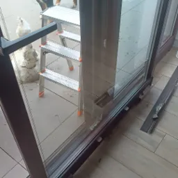 Serwisowane przesuwne drzwi tarasowe z demontowanymi elementami, drabina i pies Berneński w tle. Widoczne ślady naprawy i regulacji.